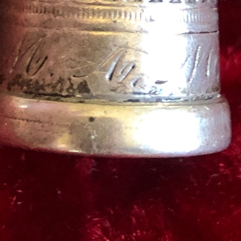 May include: Close-up of a silver thimble with engraved text. The thimble has a rounded base and a textured top edge. The text appears to be initials or a name. The background is a deep red.