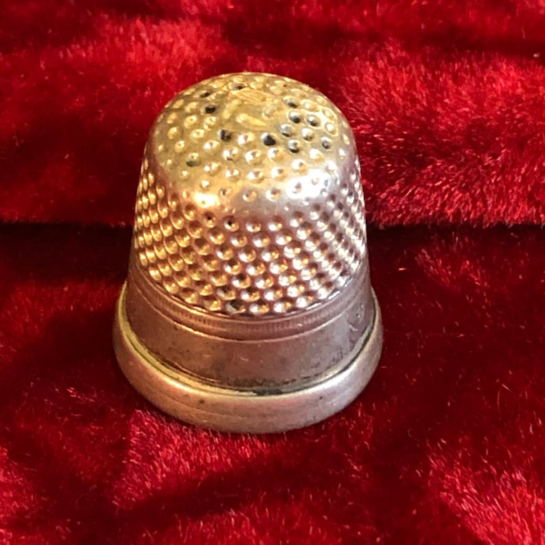 May include: A silver thimble with a rounded top and a series of small, evenly spaced holes. The thimble has a slightly flared base and is set against a deep red, textured background.