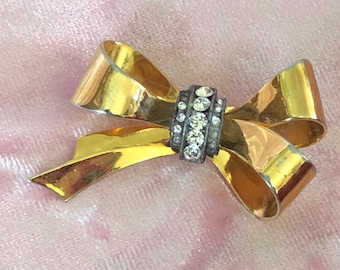 May include: A gold-toned bow brooch with a central rectangular accent. The accent is silver-toned and encrusted with clear, sparkling rhinestones. The bow's loops and tails are smooth and reflective, creating a classic, elegant accessory.