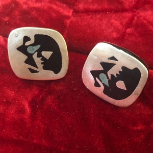 May include: A pair of square silver earrings with a black and turquoise abstract design. The earrings have a brushed metal finish and a geometric pattern. The design features a face-like shape.