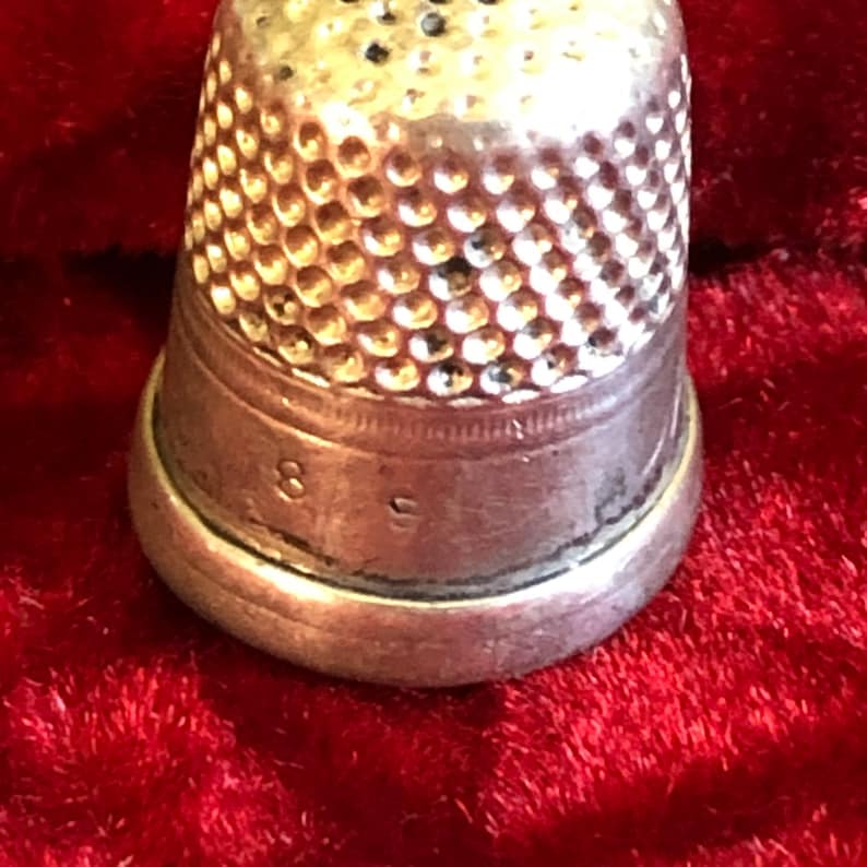 May include: Close-up of a silver thimble with a textured, dimpled surface. The thimble has a rounded top and a slightly flared base. The number "8" and the letter "S" are stamped on the base. The thimble is a sewing tool.