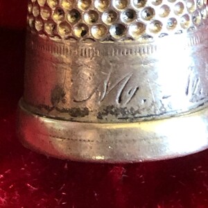 May include: Close-up of a silver thimble with a textured top and engraved markings. The thimble has a slightly tarnished appearance, with visible wear and tear. The base of the thimble is smooth and rounded.