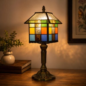 May include: A stained glass table lamp with a colorful, geometric shade. The shade features squares of blue, orange, yellow, and green. The lamp has a bronze-colored base and stem, and sits on a wooden surface.