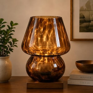May include: A decorative table lamp with a brown, textured glass shade. The lamp has a rounded base and a flared top, sitting on a wooden base. The lamp is lit, casting a warm glow. The lamp is next to a small plant in a vase.