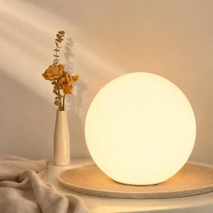 Może przedstawiać: Kulista lampa stołowa emitująca miękkie, rozproszone światło. Lampa stoi na okrągłej, beżowej tacy. Po lewej stronie znajduje się mały biały wazon z suszonymi kwiatami. Całość ma minimalistyczną i ciepłą estetykę.