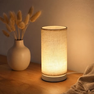 May include: A cylindrical table lamp with a beige linen shade and a white base. The lamp is illuminated, casting a warm glow. A vase with dried flowers sits to the left. The lamp is on a wooden surface.