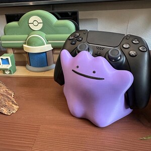 以下が含まれることがあります： 紫色のDitto型のコントローラーホルダーと黒いコントローラー。ホルダーには笑顔があります。背景には、緑と白のポケモンをテーマにしたディスプレイがあります。