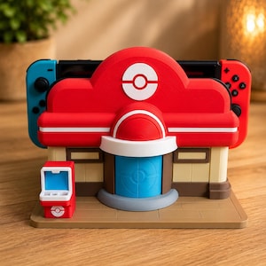 May include: A red, white, and blue plastic gaming console dock shaped like a building with a circular logo on the roof. The structure features a central blue door, a small red side kiosk, and a tan base, designed to hold a handheld gaming device.