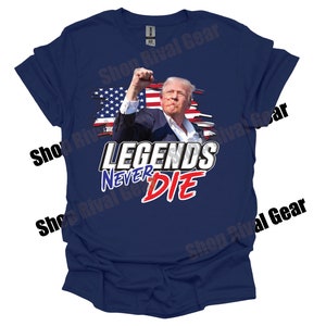 May include: A navy blue t-shirt featuring a distressed American flag design and the text "Legends Never Die" in white and red lettering. The image depicts a man in a suit with his fist raised in the air.