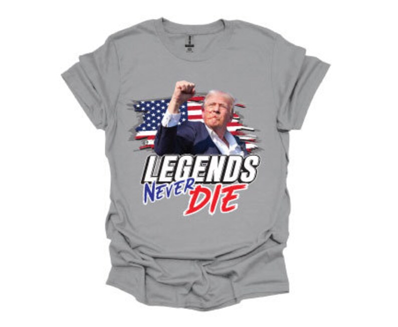 May include: A grey t-shirt featuring a graphic of a man in front of an American flag with the text "Legends Never Die" in red, white and blue.