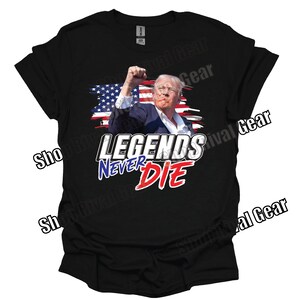 May include: Black t-shirt featuring a graphic of a man with his fist raised in the air. The graphic is in front of a red, white and blue American flag. The text "LEGENDS NEVER DIE" is printed in white, red and blue lettering below the graphic.
