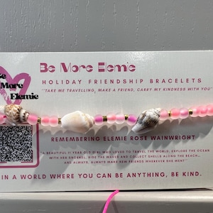May include: A pink beaded bracelet featuring three seashells and gold-coloured accents. The bracelet is presented on a card with the text "Be More Elemie" and "Holiday Friendship Bracelets". The card also includes a QR code and text about Elemie.