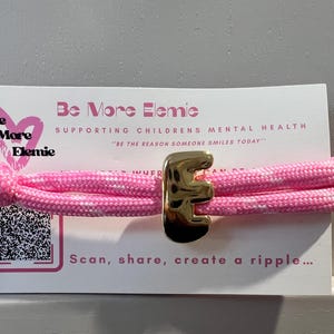 May include: A pink and white bracelet with a gold letter "E" charm. The bracelet is displayed on a card with the text "Be More Elemie" and "Supporting Children's Mental Health". The card also includes a QR code and the phrase "Scan, share, create a ripple..."