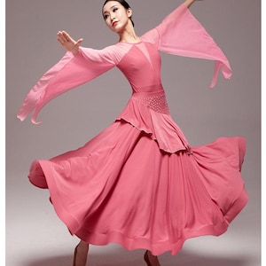 Peut inclure: Une robe de danse de salon rose avec de longues manches transparentes et une jupe à volants. La robe présente un corsage ajusté et une jupe évasée, créant une silhouette dynamique. La danseuse porte des talons bruns.