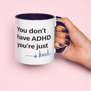 May include: White ceramic mug with a navy blue interior and handle. The mug features the text "You don't have ADHD you're just a knob" in black and blue lettering. The mug is held by a hand with blue nail polish.