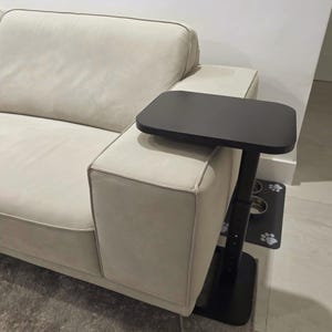 May include: A black, adjustable side table with a rectangular tabletop is positioned next to a beige sofa. The table has a vertical support and a base. The sofa has a modern design with a light-colored fabric.