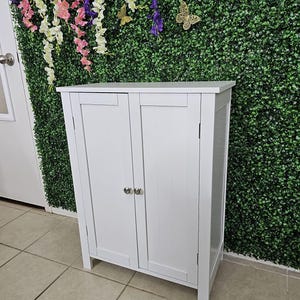 May include: A white wooden cabinet with two doors and silver knobs. The cabinet is rectangular and stands on short legs. The background features a green wall with decorative butterflies and hanging flowers.