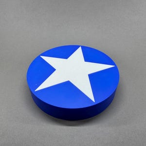 Peut inclure: Un objet rond bleu avec une étoile blanche au centre. L'étoile est un design à cinq branches. L'objet est posé sur une surface grise.
