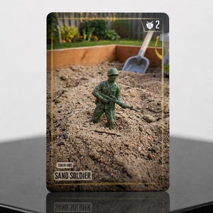 May include: A rectangular game card featuring a green plastic toy soldier standing in a sandbox. The card displays the text TOKEN UNIT SAND SOLDIER at the bottom and a shield icon with the number 2 in the top right corner, framed by a gold border.