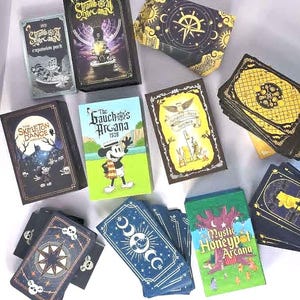 Puede incluir: Una colección de mazos de cartas rectangulares con varios temas ilustrados, incluyendo Steamboat Arcana, The Skeleton Dance Arcana, The Gaucho Arcana, Hundred-Acre Arcana y The Mystic Honeypot Arcana, con diversas paletas de colores y diseños gráficos.