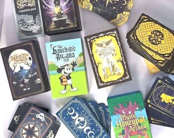 Può includere: Una collezione di mazzi di carte rettangolari con vari temi illustrati, tra cui Steamboat Arcana, The Skeleton Dance Arcana, The Gaucho Arcana, Hundred-Acre Arcana e The Mystic Honeypot Arcana, con diverse tavolozze di colori e design grafici.