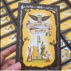May include: A deck of cards featuring a whimsical illustration with the text "Hundred-Acre Arcana." The artwork includes an owl, Winnie the Pooh, Tigger, Piglet, and Rabbit. The card has a yellow background with a brown border.