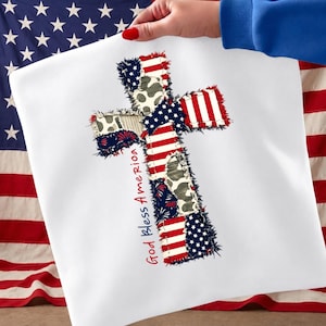 Può includere: Un tessuto quadrato bianco con un motivo a croce. La croce è composta da toppe con motivi della bandiera americana, camouflage e strisce rosse e bianche. Il testo "God Bless America" è scritto in rosso.