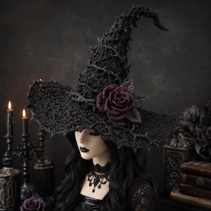 May include: A black crocheted witch hat featuring a pointed, textured crown and a wide, jagged-edged brim. A deep burgundy crocheted rose with dark green leaves is attached to the side of the hat, adding a contrasting floral element to the dark design.