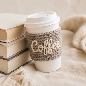 May include: A white disposable coffee cup featuring a beige crocheted sleeve with the word Coffee embroidered in white yarn. The cup is positioned next to a stack of three books with light-colored covers, all resting on a textured fabric surface.