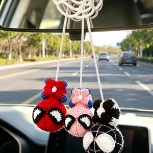 May include: Three crocheted spherical ornaments hanging from a white circular crocheted base. The ornaments feature red, light pink, and black yarn colors with white web-like patterns and large white eye shapes, suspended by white strings.