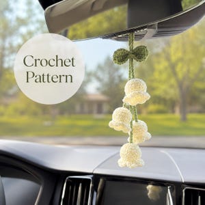 May include: A hanging crochet ornament featuring four cream-coloured bell-shaped flowers attached to a green stem with a small green leaf bow at the top. The text Crochet Pattern is visible on the left side of the image, suspended from a mirror.