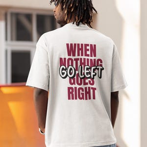 Op de afbeelding: Een lichtgrijs t-shirt met korte mouwen en de bordeauxrode tekst WHEN NOTHING GOES RIGHT en de zwarte tekst GO LEFT op de achterkant. Het shirt wordt gecombineerd met een lichtgrijze baseballpet en een blauwe spijkerbroek, gezien vanaf de rug.