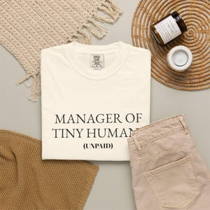 Op de afbeelding: Crèmekleurig T-shirt met de tekst "MANAGER OF TINY HUMAN (UNPAID)". Het shirt wordt gecombineerd met een beige broek, een geweven placemat, een kaars en een decoratief kleed. Het shirt heeft een Comfort Colors label.