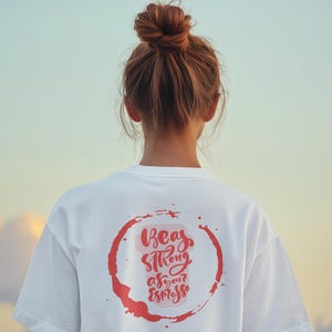 Op de afbeelding: Een wit t-shirt met een rode cirkelvormige koffievlek-afbeelding op de achterkant. Binnen de cirkel staat de tekst "Beas strong as your Espresso" in een gestileerd lettertype, met de woorden "Espresso Appreciation Society" onder de afbeelding.