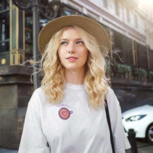 Op de afbeelding: Een wit T-shirt met korte mouwen en een kleine afbeelding op de borst van een paarse koffiekop met een hartje erin en de tekst "MORNING CLUB" erboven gedrukt, gedragen met een beige strooien hoed met een brede rand.