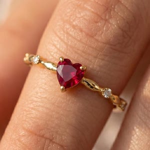 Op de afbeelding: Een gouden ring met een hartvormige rode edelsteen. De band heeft een delicaat ontwerp met kleine, heldere accentstenen. De ring wordt getoond op een vinger, wat de elegante en romantische stijl benadrukt.