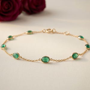 May include: A delicate gold bracelet featuring oval-cut emeralds. The bracelet is strung with small gold links and has a clasp closure. Two red roses are in the background. The bracelet is a piece of fine jewelry.
