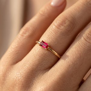 Op de afbeelding: Een gouden ring met een rechthoekige, helderrode edelsteen. De ring heeft een slanke, gepolijste gouden band en de edelsteen is horizontaal gezet. Het ontwerp is eenvoudig en elegant, geschikt voor dagelijks gebruik of speciale gelegenheden.
