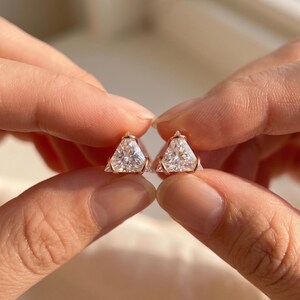 May include: A pair of stud earrings featuring triangular-cut clear stones set in rose gold metal. Each earring has a three-prong setting that secures the faceted stone, creating a geometric shape that reflects light across the surface.
