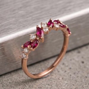 May include: A rose gold ring featuring a curved arrangement of alternating marquise-cut red rubies and round-cut white diamonds. The band is thin and polished, showcasing a delicate design where the gemstones are set in a continuous, flowing pattern.