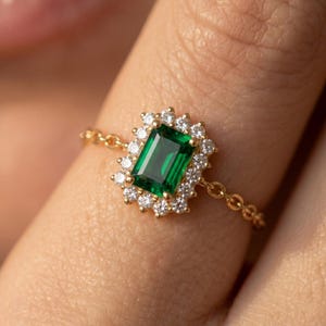 Könnte beinhalten: Ein goldener Ring mit einem smaragdgrünen, rechteckig geschliffenen Edelstein, umgeben von einem Kranz aus kleineren, klaren Edelsteinen. Der Ring hat ein filigranes Kettendesign am Band. Der Ring wird an einem Finger getragen.