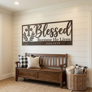 Puede incluir: Un letrero de pared rectangular de metal marrón oscuro con un icono de cruz recortado, dos ramas con hojas y el texto Blessed Because He Lives John 14:19 en una combinación de fuente cursiva y serif, montado en una pared blanca.