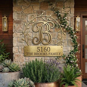 May include: A gold-toned metal wall sign featuring a large script letter B enclosed in a circular vine frame, mounted above a rectangular plaque with the text 5160 and THE BROOKS FAMILY, displayed on a textured stone wall with two lanterns.