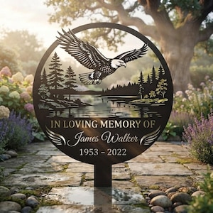 May include: A circular black metal memorial plaque featuring a laser-cut design of a bald eagle in flight over a forest and lake. The text reads IN LOVING MEMORY OF James Walker 1953 - 2022. The plaque is mounted on a stake for ground placement.