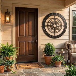 May include: A circular, black metal compass rose sign with the text "HOME IS HERE THE WILLIAMS" is mounted on a wooden wall. A brown wooden door is to the left. Potted plants and a welcome mat are in front.