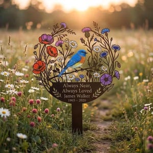 May include: A circular metal garden stake featuring a bluebird perched on a branch surrounded by colorful poppies, daisies, and thistles. The text reads: Always Near, Always Loved, James Walker, 1965-2020. The design is dark brown metal.