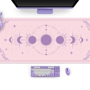 May include: A pink desk mat with a celestial design in purple, featuring moons, suns, and stars. A matching purple keyboard, mouse, and headset with cat ears are also visible, along with a purple computer monitor.