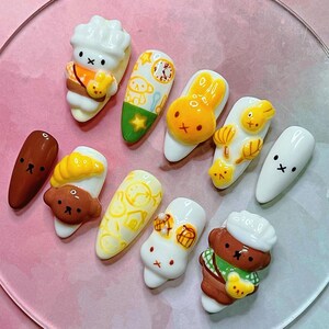 May include: A collection of whimsical nail art designs featuring cartoon characters and patterns. The nails showcase a variety of colors, including brown, yellow, white, and orange. Each nail has a unique design, such as a bear, bunny, or clock.