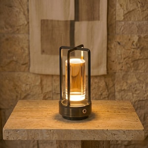May include: A contemporary lamp with a cylindrical glass shade, emitting a warm light. The lamp is enclosed in a dark metal frame and has a bronze-coloured base. It sits on a stone table. The overall design is minimalist.