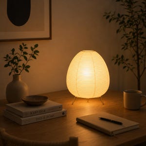 May include: An illuminated, egg-shaped table lamp with a paper shade and three metal legs. The lamp is on a wooden desk alongside books, a mug, and a notebook. The warm light creates a cosy atmosphere.